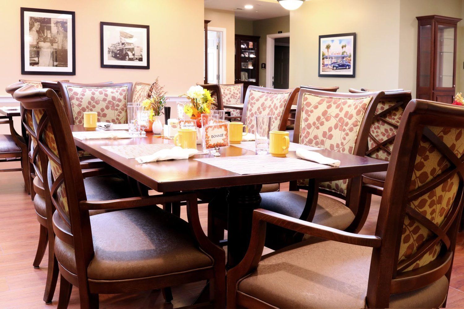Dining room with a long wooden table set with placemats and chairs. Paintings on the wall.