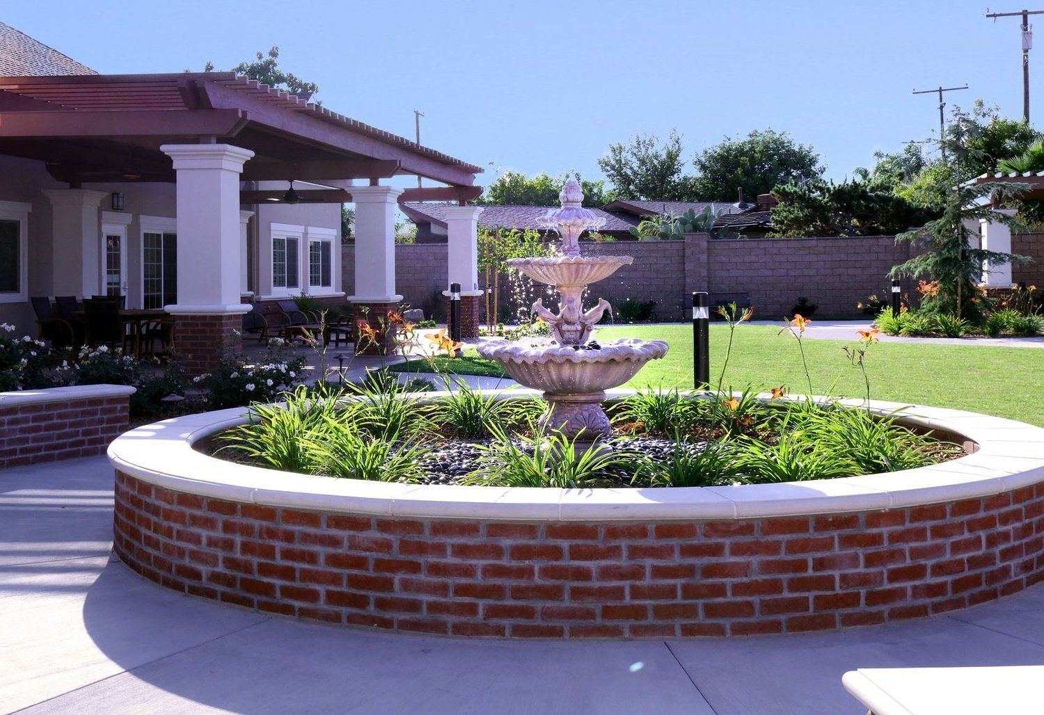 Fountain in a circular brick planter, in front of a building with columns and red roof. Green grass and bushes surround.