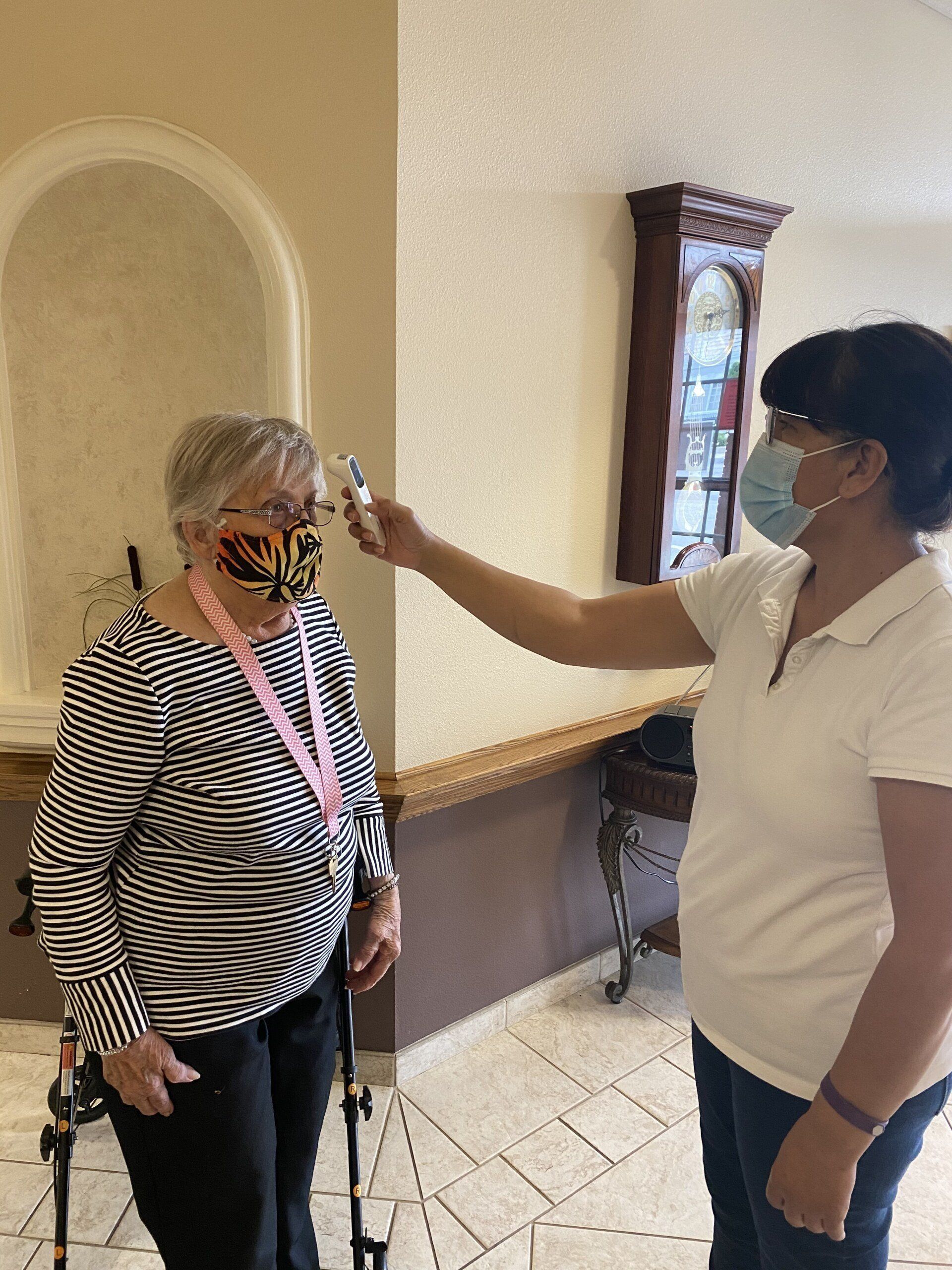 A healthcare worker checks a woman's temperature with a thermometer. Both are wearing masks, indoors.