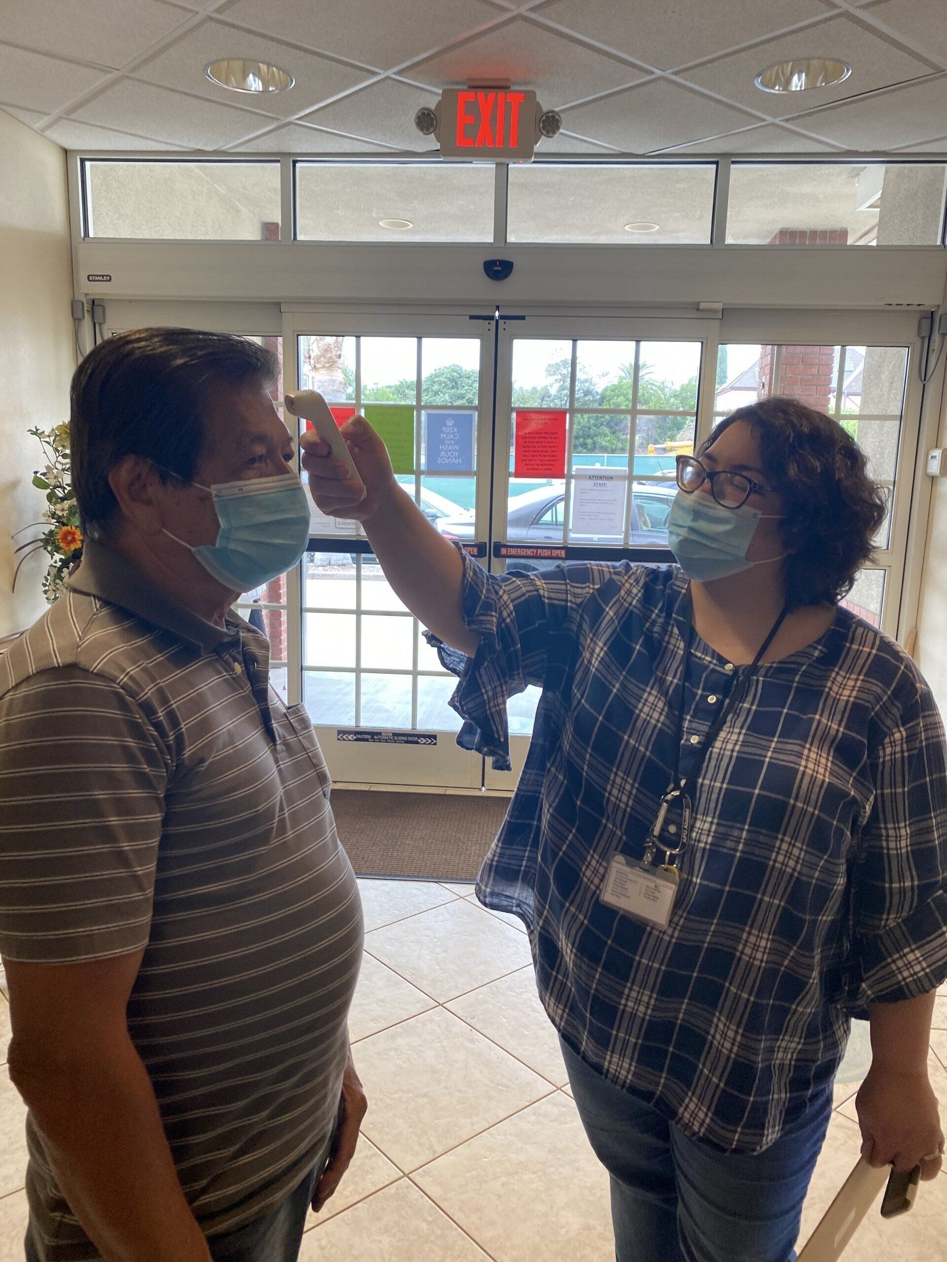 Woman taking man's temperature with thermometer at building entrance; both wearing masks.