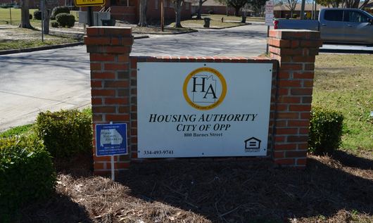 Sign for Housing Authority of Opp, City of Opp. White sign with logo between brick columns, shrubbery in front.