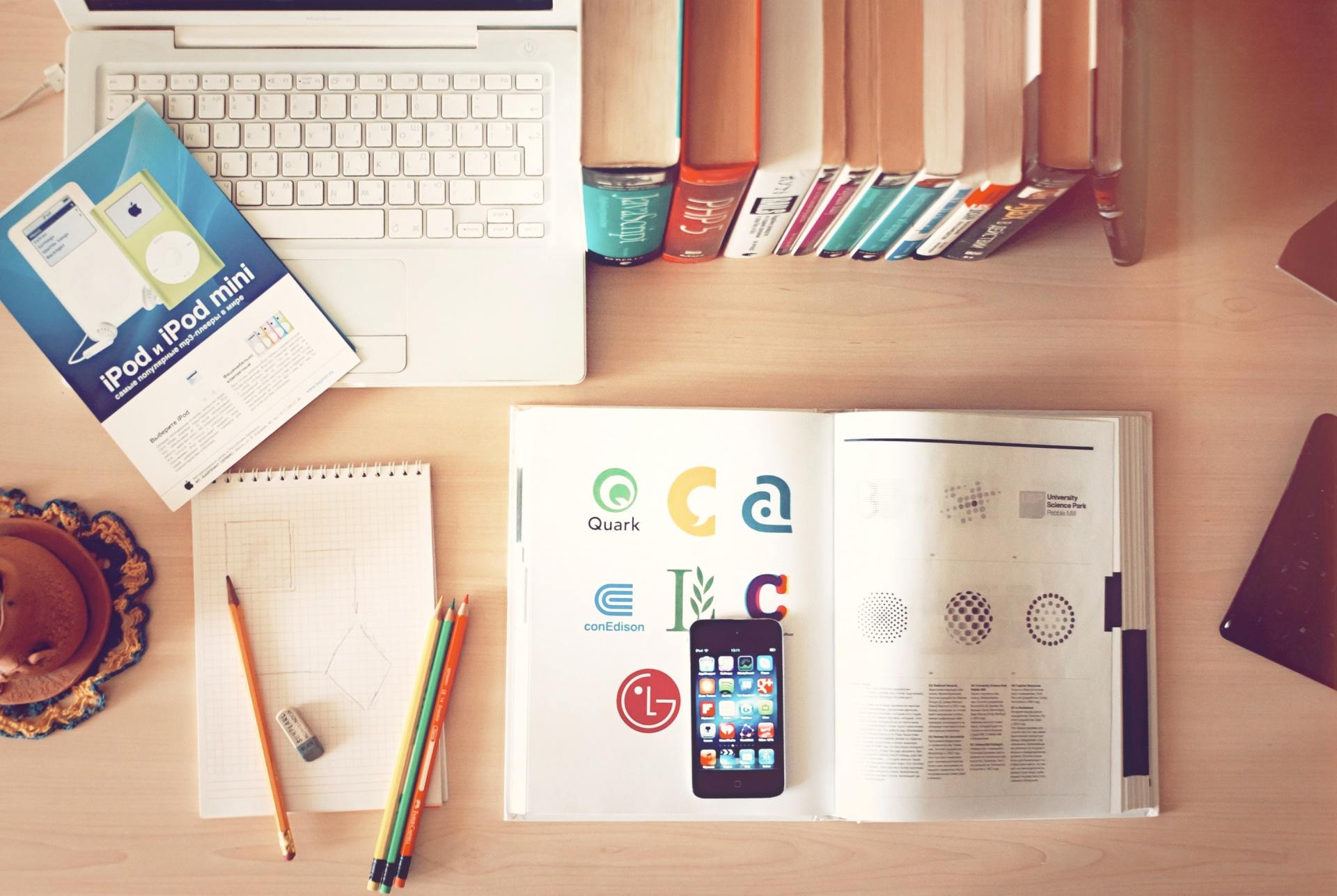 Laptop, open book with logos, iPod, pencils, and books on a wooden desk.