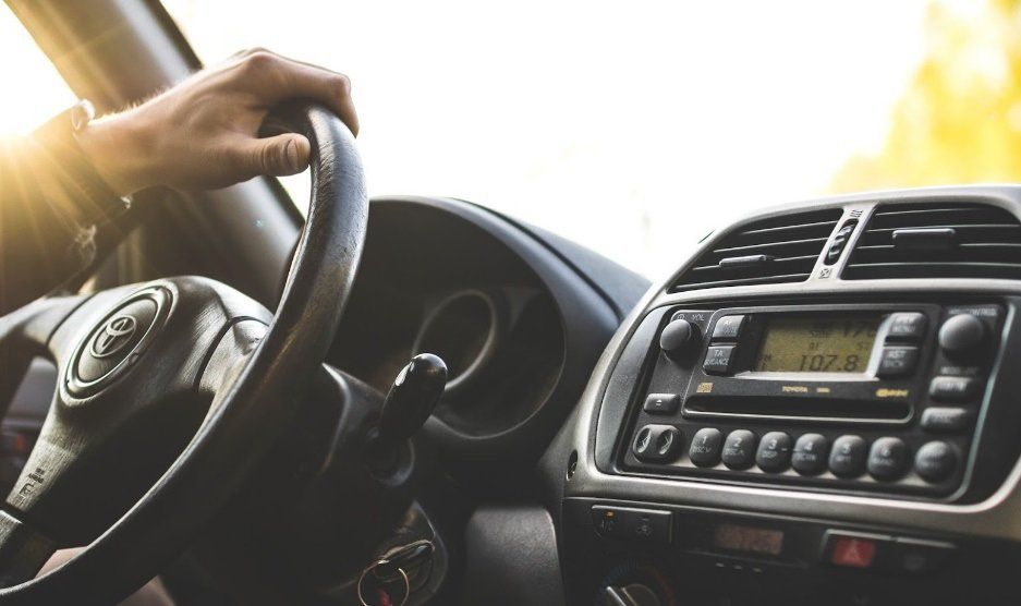 Hand on steering wheel inside a car, dashboard visible; bright sunlight.