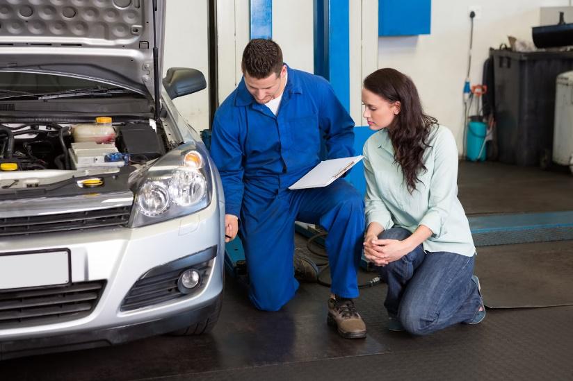 Mechanic in blue jumpsuit shows car owner paperwork at a car repair shop.