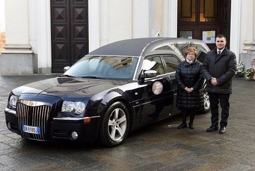 Una donna e un uomo sono in piedi accanto a un carro funebre Chrysler nero davanti a un edificio con grandi portoni.