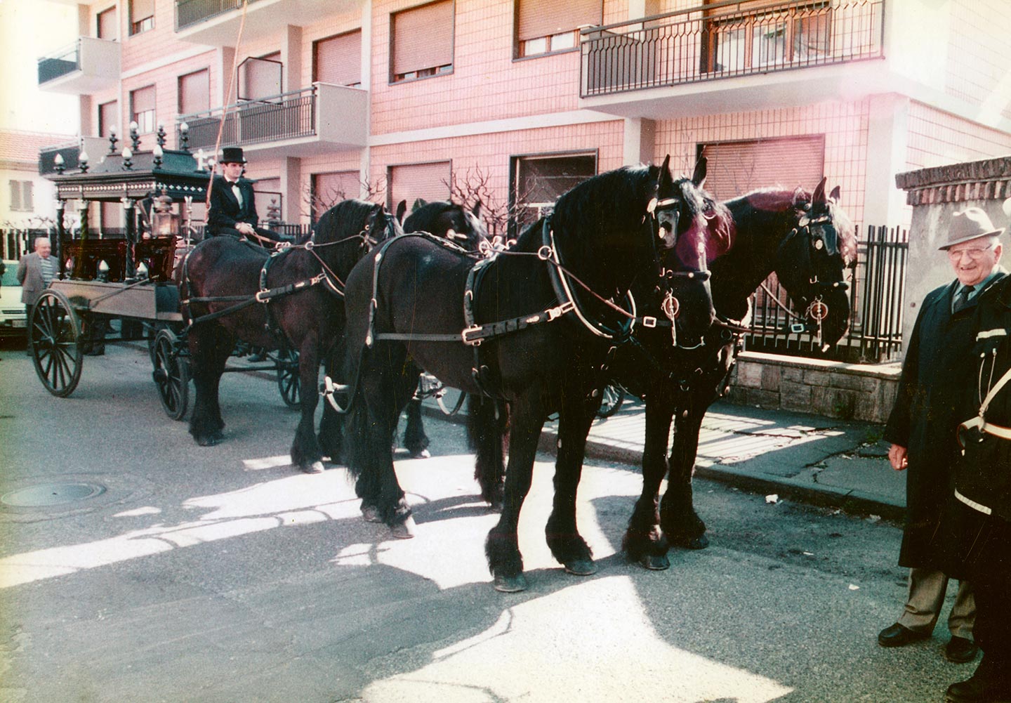 Una tradizionale carrozza funebre trainata da quattro cavalli neri è ferma in strada, accanto a un uomo in abito formale.