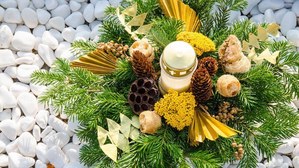 Una composizione funeraria festiva con una candela centrale, rametti di sempreverdi, pigne e dettagli dorati su pietre bianche.