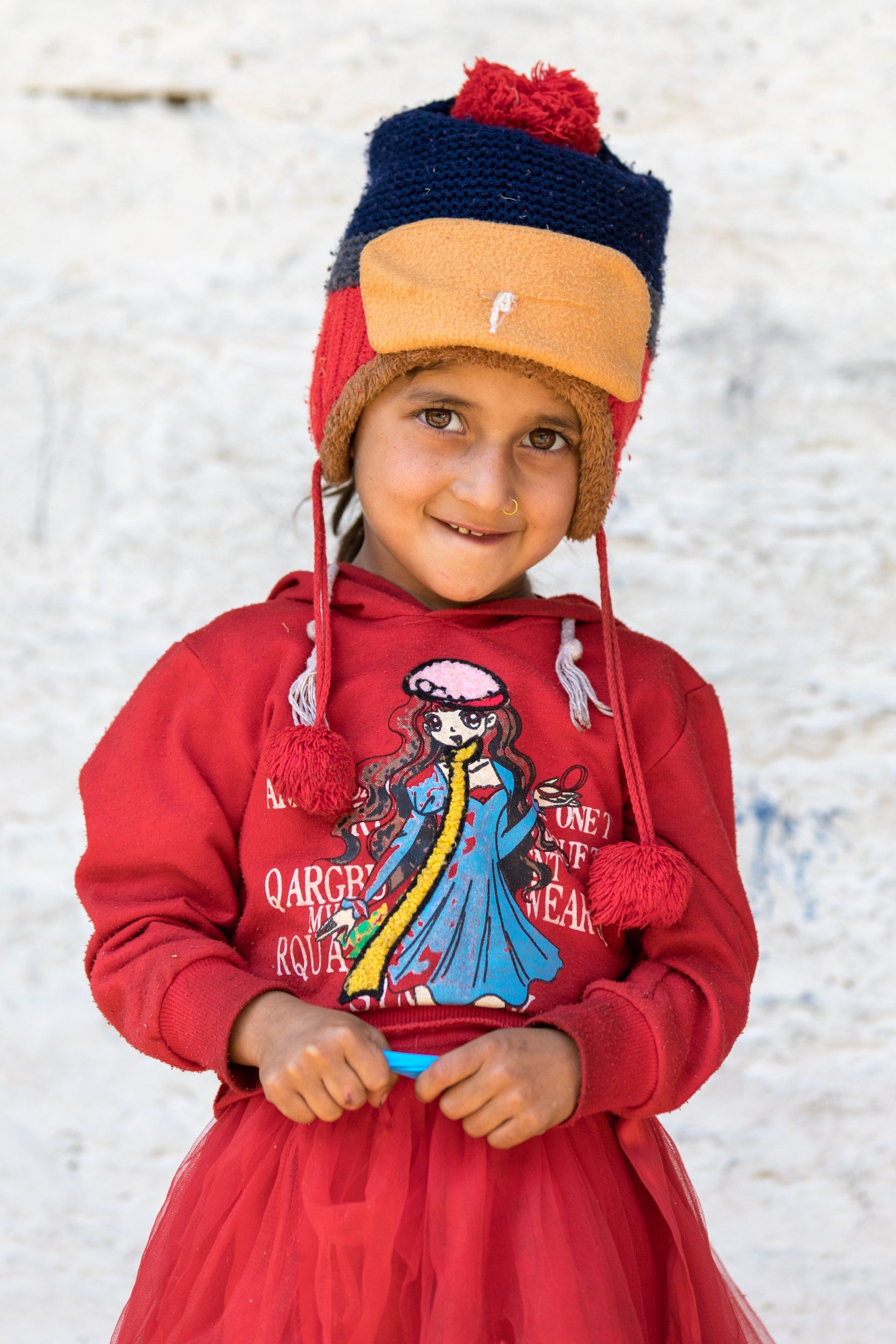 A little girl wearing a red dress and a hat is standing in the snow.