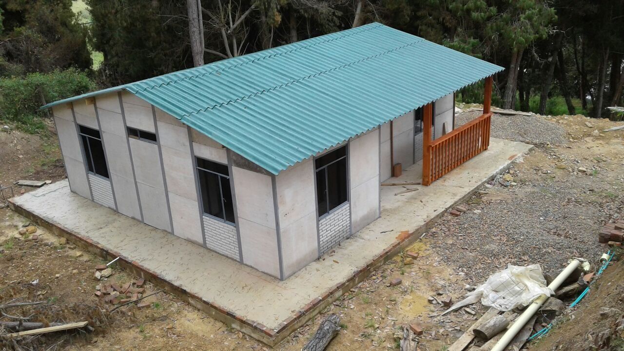 Pequeña casa de una sola planta con techo de metal color turquesa, situada en un terreno de grava y rodeada de árboles.