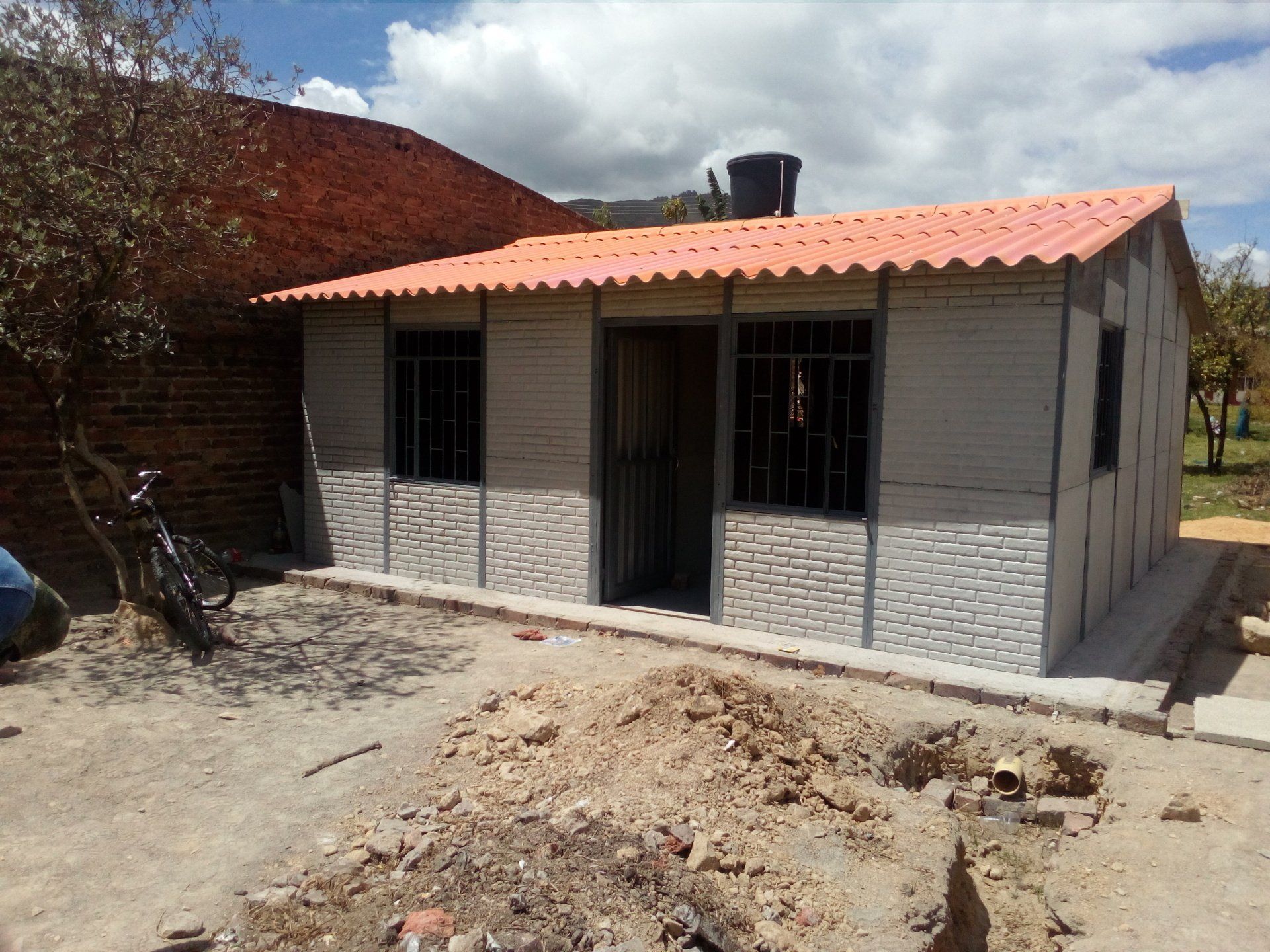 Pequeña casa gris con techo rojo en un patio seco, vista desde el frente.