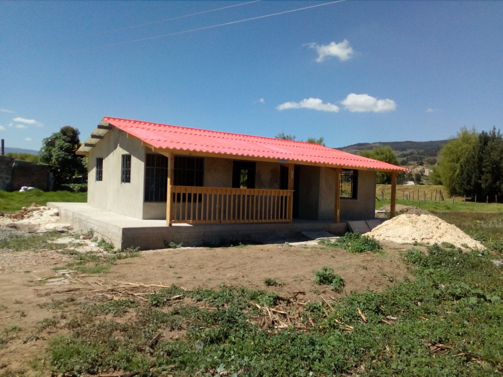 Pequeña casa color crema con techo rojo y porche en un campo rural bajo un cielo azul.