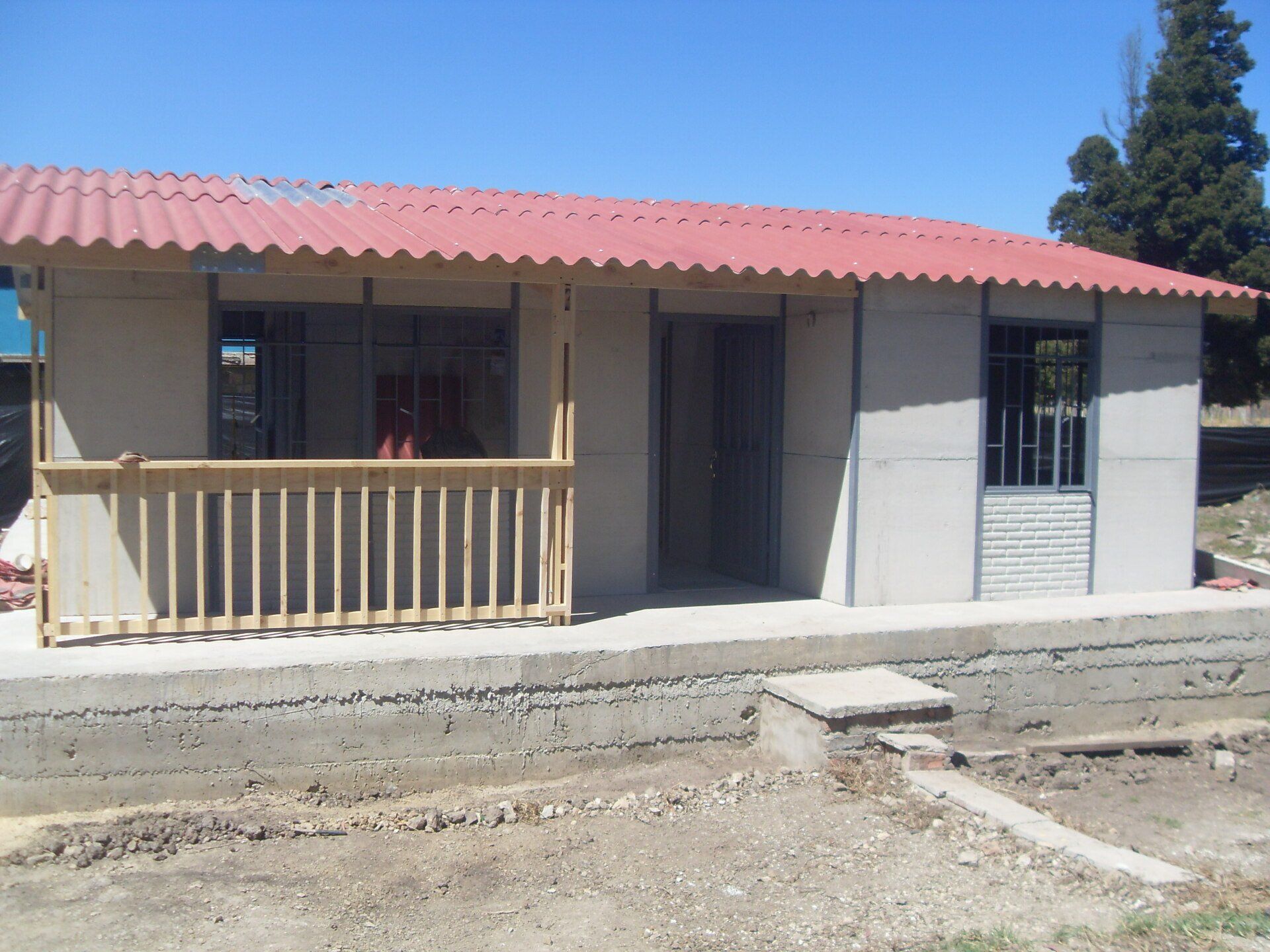 Pequeña casa de una sola planta con techo rojo, porche delantero y ventanas enrejadas bajo un cielo azul claro.