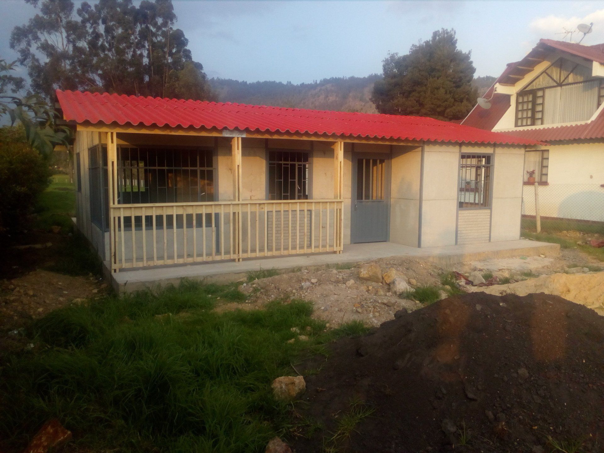 Pequeña casa color crema con techo y porche rojos, situada en un patio rural cubierto de hierba al atardecer.