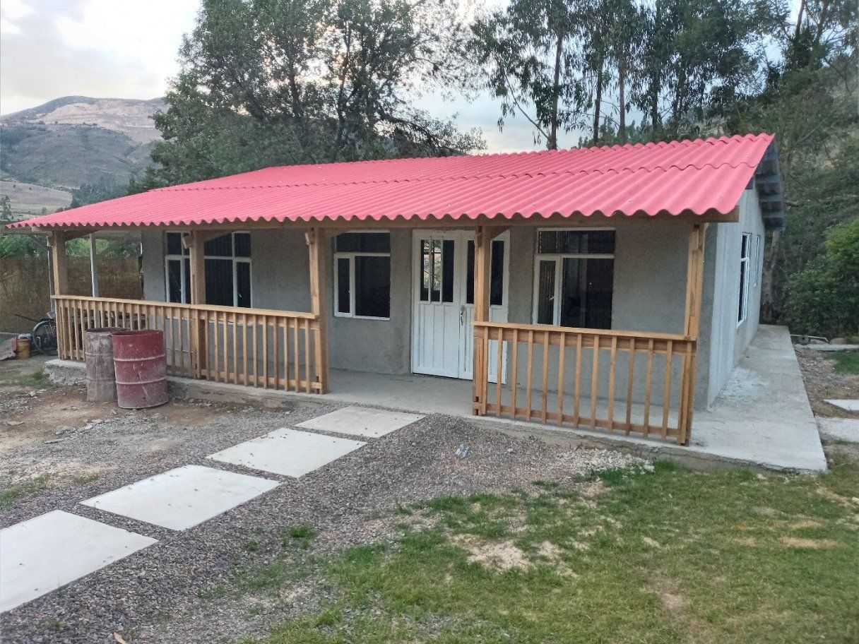 Pequeña casa gris con techo rojo, porche delantero y camino de acceso en un patio rural.