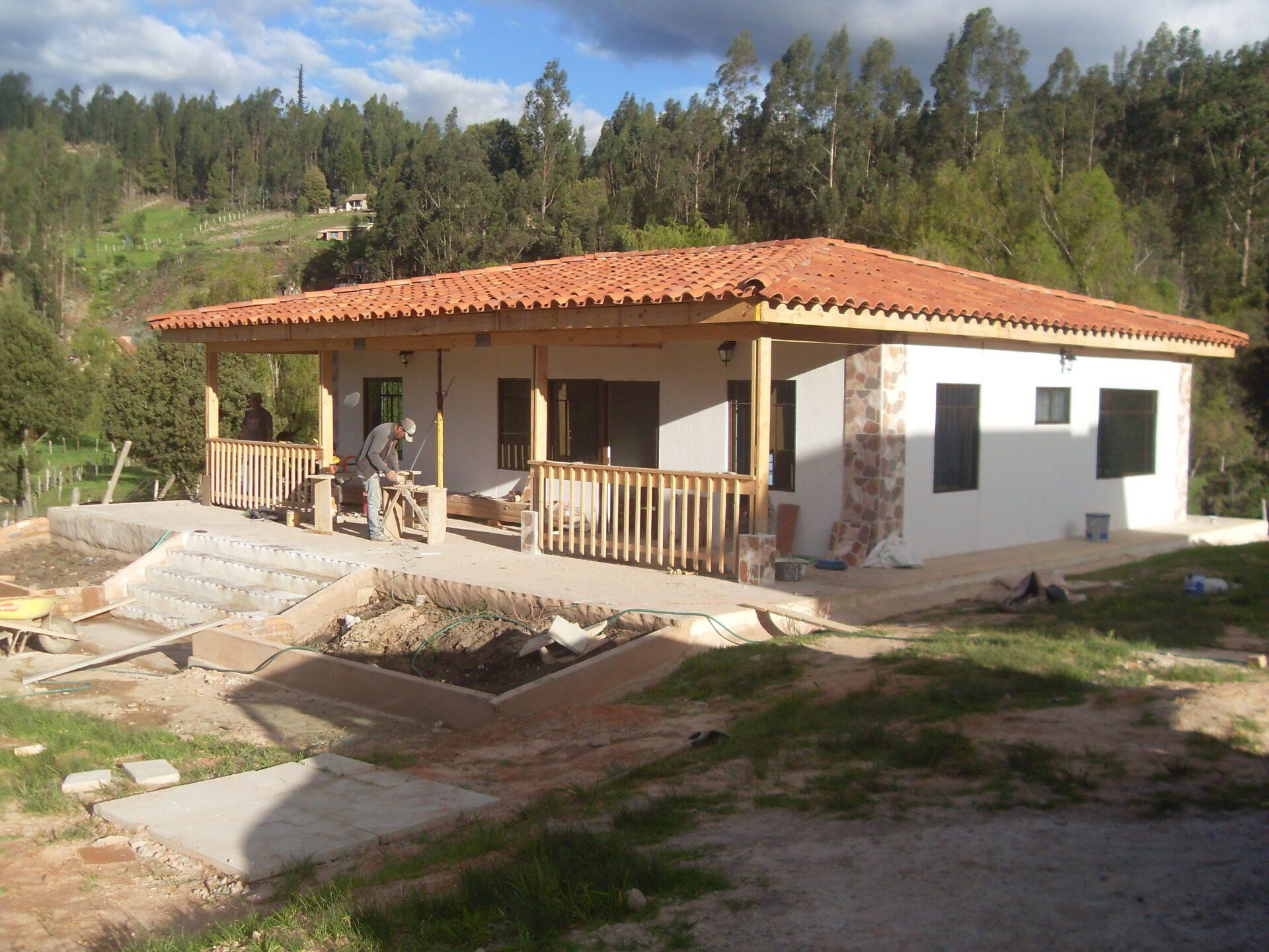 Pequeña casa blanca con techo de terracota y porche en una zona rural y boscosa.