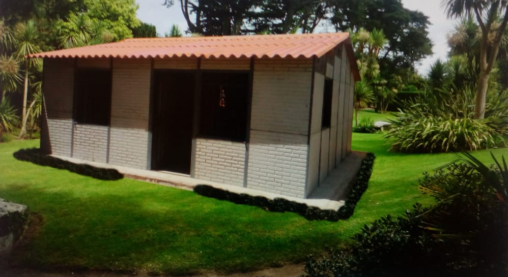 Pequeña casita de color beige con techo rojo sobre un césped verde, rodeada de árboles y plantas de jardín.