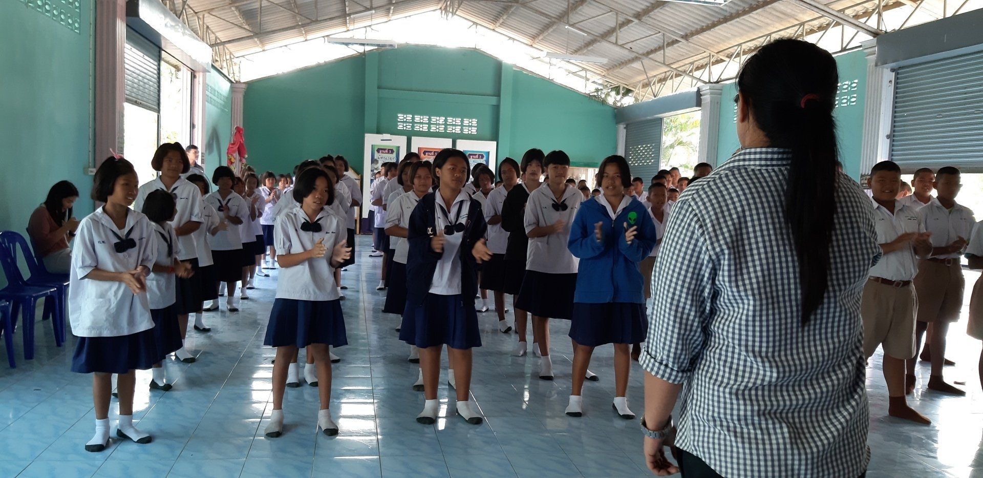 Students in uniforms standing in a room, led by a person in a plaid shirt, possibly doing exercises.