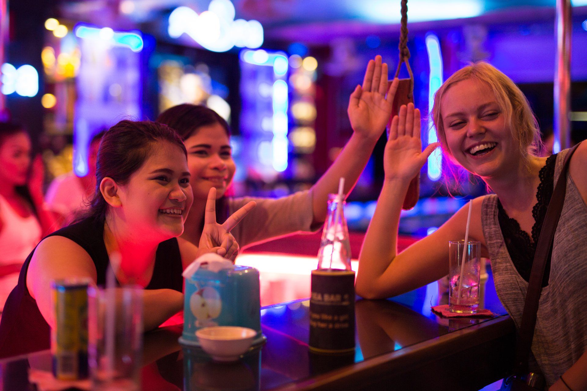 Three people at a bar waving and smiling, neon lights in background.