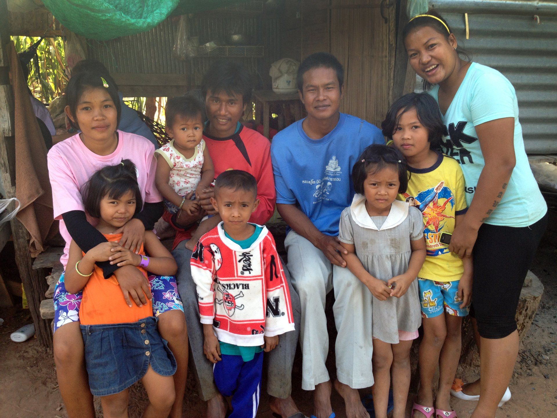 Family of nine poses outdoors, smiling. Children wear colorful clothes; adults wear casual attire, near a wooden structure.