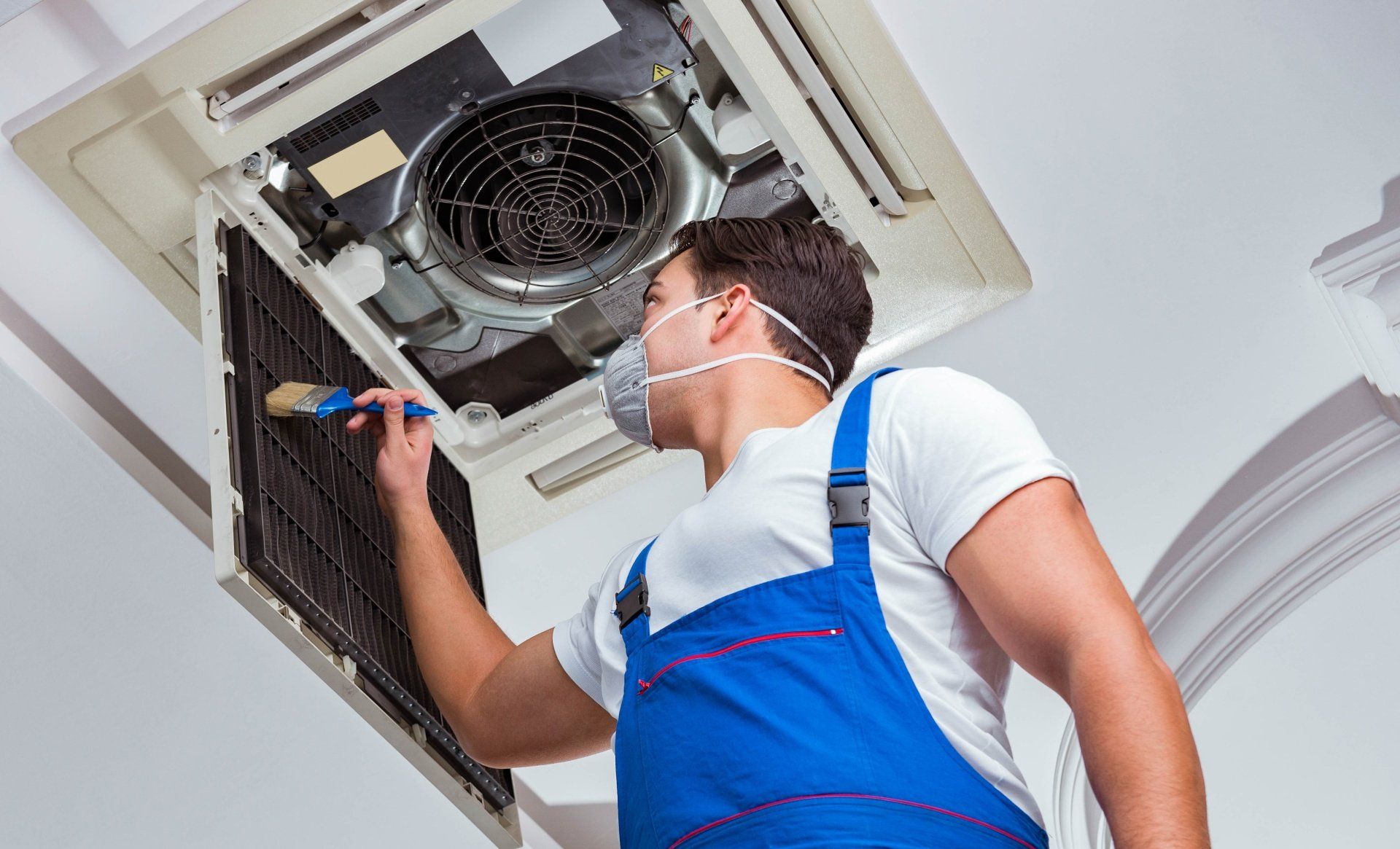 A man wearing a mask is cleaning an air conditioner with a brush.
