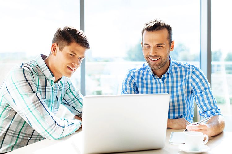 Two men are sitting at a table looking at a laptop computer.