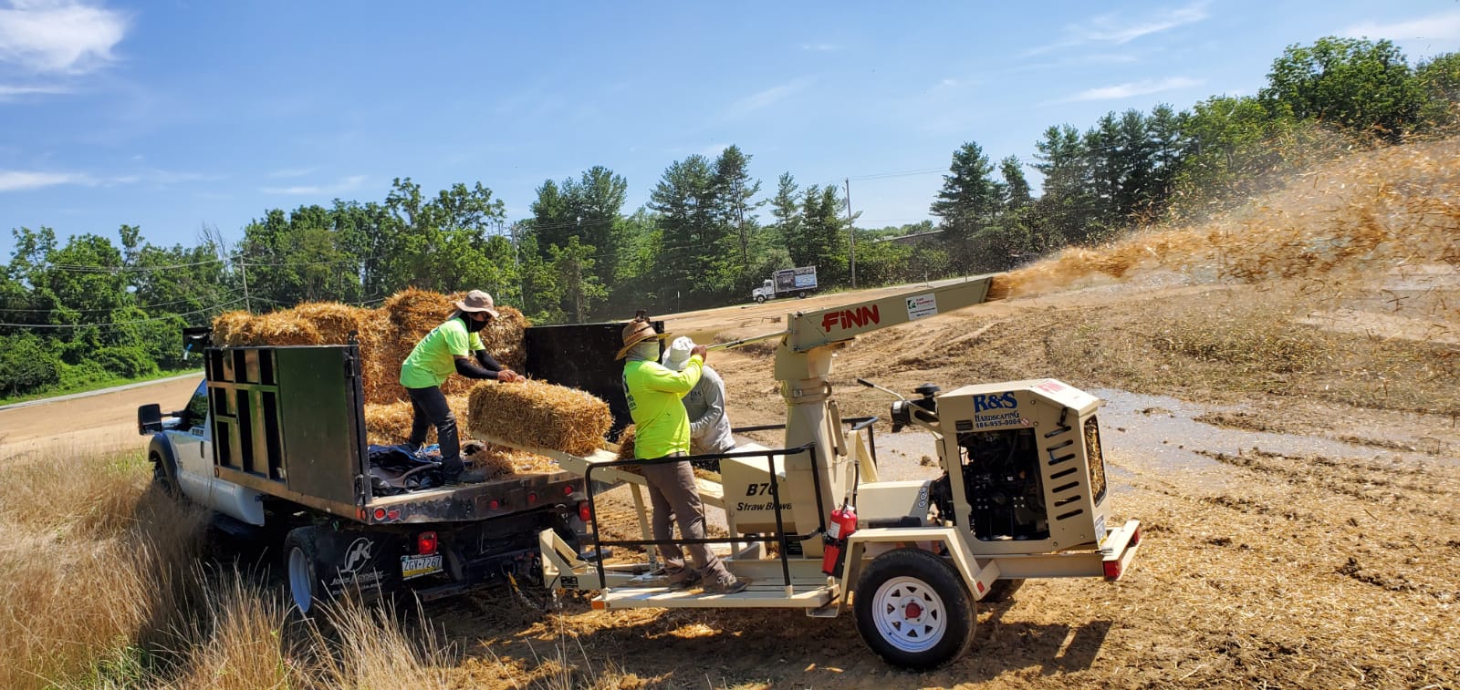 Workers feeding hay bales into a wood chipper. Chipped hay blows into a large pile. Sunny day.