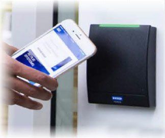Hand holding a smartphone near a black access control reader; screen shows a company app.