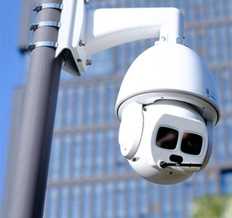 White security camera mounted on a pole, overlooking a building with blue-tinted windows.