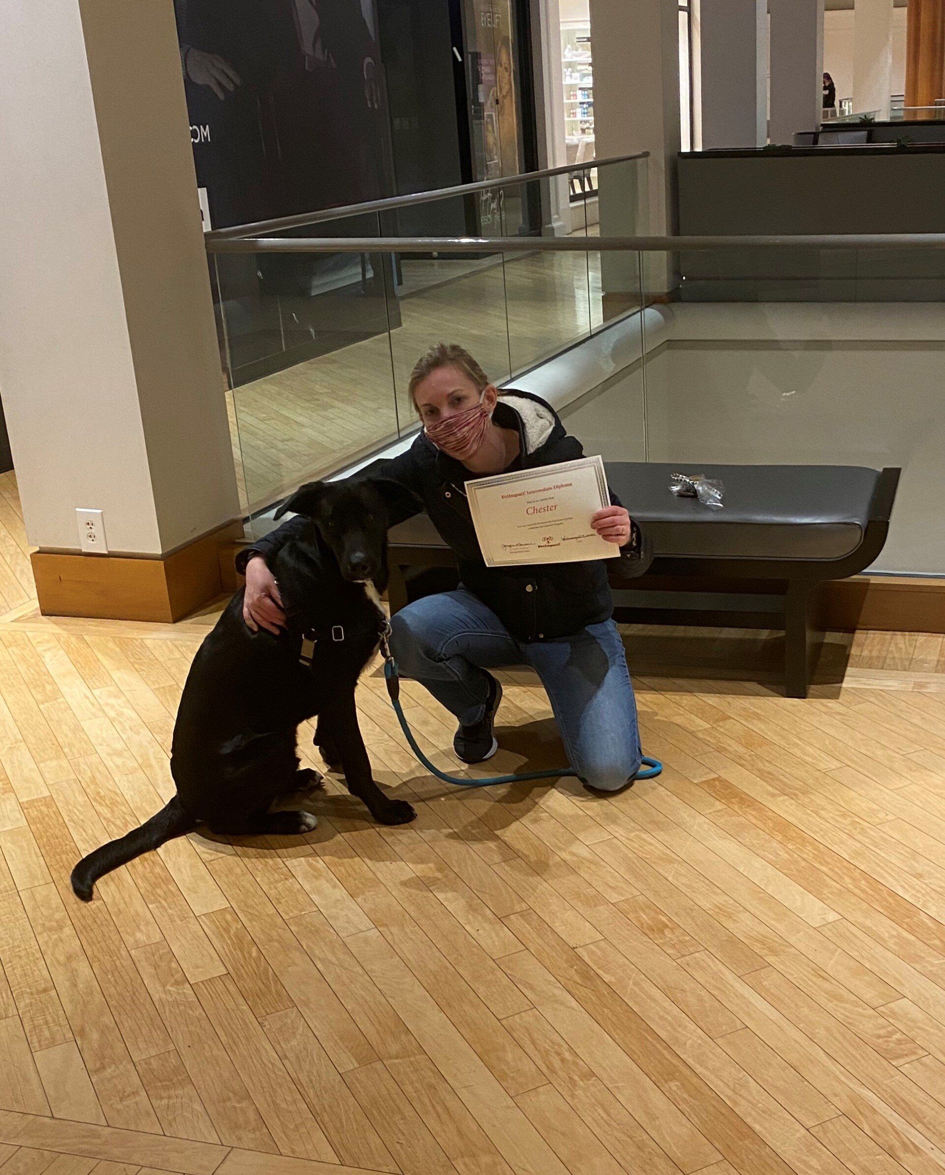 Woman kneeling next to a black dog in a building, holding a paper; wooden floor and railing visible.