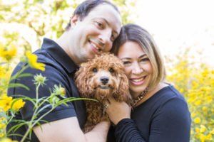 Couple smiling, holding a small brown dog in a field of yellow flowers.