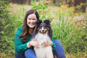 Woman smiling, hugging a tricolor Sheltie dog in a garden with green foliage and flowers.