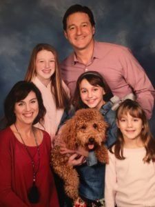 Family of five, including a dog, smiling in a portrait. Man in pink shirt, woman in red, girls in light tops.