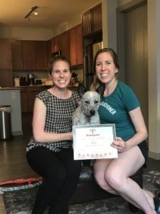 Two women, a dog, and a certificate inside a home; the women are smiling, holding the dog and certificate.