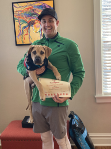 Man holding dog and certificate, smiling indoors. Dog wearing sweater. Green jacket, gray shorts.