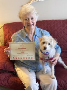 Woman and small white dog seated on a couch, holding a framed certificate. The woman smiles.