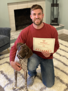 Man kneeling with dog, holding a certificate. Fireplace and rug in the background. Man smiles.