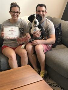 Couple and dog on a couch holding a certificate. The woman wears glasses and red shorts. The dog is black and white.