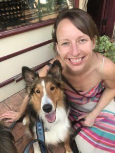 Woman smiling with a tri-color dog outdoors. They're sitting close, dog panting, woman in striped dress.