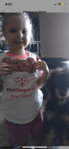 Girl holding a dog treat, smiling at the camera, with a black poodle. She wears a white