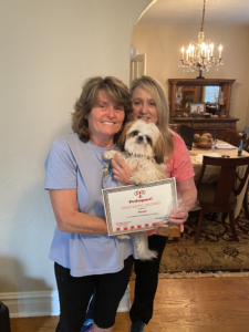 Two women holding a Shih Tzu dog and a certificate indoors. The dog wears a collar. They are smiling.