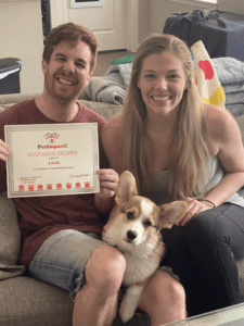A smiling couple with a corgi holds a framed certificate; they sit on a couch inside a home.