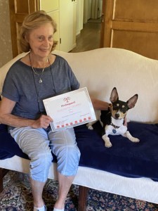 Woman and small dog on a sofa; woman holds a certificate, dog looks at the camera.