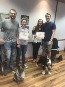 Four people with two dogs hold certificates in an office setting.