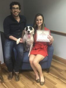 Man, woman, and dog sitting on blue chair; woman holds a framed certificate.