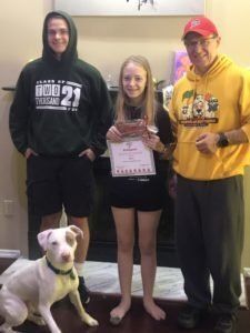 Group of three people and a dog by a fireplace; one holds a certificate.
