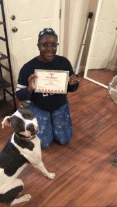 Person kneeling, holding a certificate. Dog sits next to the person. Wooden floor, room.