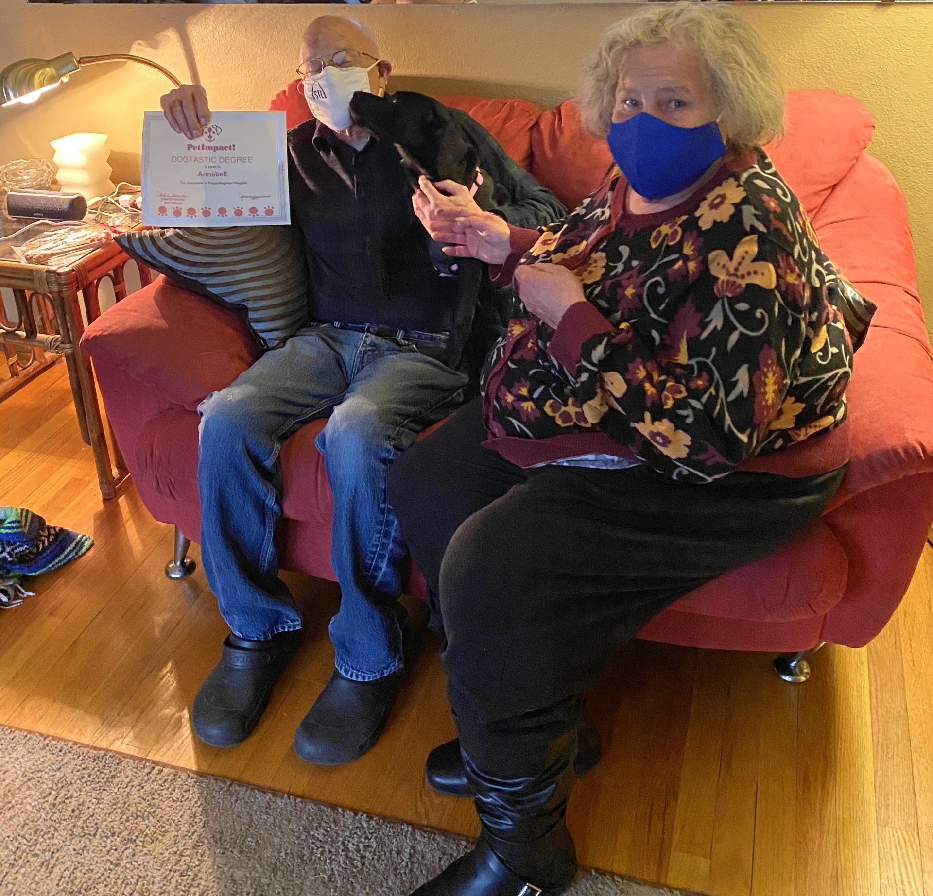 Two people wearing masks sit on a red couch, holding a certificate.