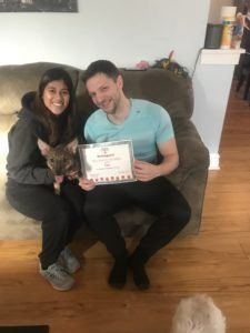 Couple sitting on a couch with a dog, holding a certificate. They are smiling indoors.