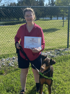 Woman holding certificate, stands with a Doberman Pinscher in front of a chain link fence.
