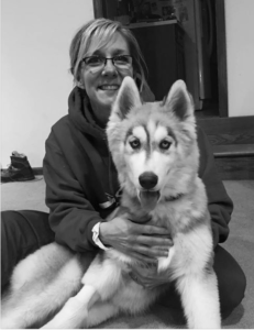 Woman in glasses with a husky dog, both smiling. They sit indoors.